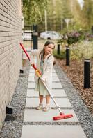 un pequeño niña con un cepillo limpia un camino en el calle en el patio foto