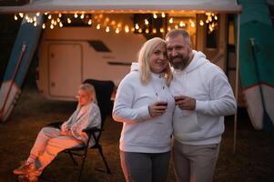 un casado Pareja con lentes de vino soportes en contra el antecedentes de un casa rodante y descansa juntos por el hoguera. noche familia vacaciones foto