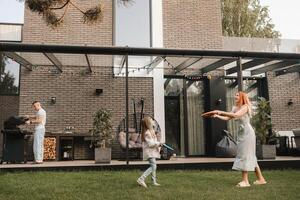 A happy family is resting near their home. dad is cooking a barbecue, and mom and daughter are playing photo