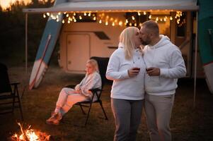 A married couple with glasses of wine stands against the background of a motorhome and rests together by the campfire. Evening family vacation photo