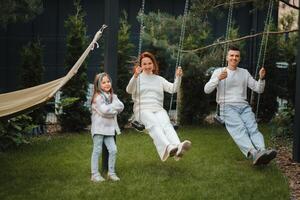 Mom and dad are riding on a swing and there is a daughter standing next to them. The family is resting on a swing photo