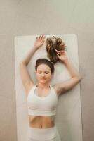 un niña en blanco ropa lo hace yoga acostado en un alfombra adentro foto