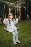 A beautiful girl with long hair in a jacket rides on a swing in the evening on the street photo