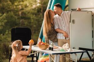 un joven familia es descansando siguiente a su móvil hogar. el niña toma un naranja desde el mesa, mientras el padres hacer no ver foto