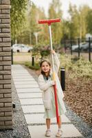 un pequeño niña con un cepillo limpia un camino en el calle en el patio foto