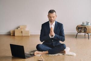 A man in a formal suit meditates while sitting in a fitness room with a laptop photo