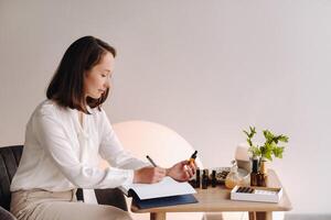 The aromatherapist girl is sitting in her office and holding a bottle of aromatic oil in her hands and writing something down. there are essential oils on the table photo