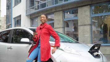 Afrikaanse vrouw staat met smartphone in de buurt haar elektrisch auto en looks Bij de camera. opladen elektrisch auto Bij de elektrisch gas- station video