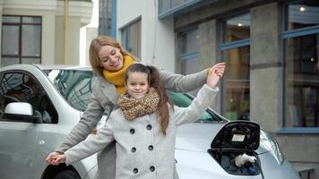 een vrouw met haar dochter staat in de buurt haar elektrisch auto en looks Bij de camera. opladen een elektrisch auto Bij een gas- station video