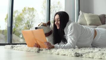 giovane donna è lettura libro e carezzevole sua animale domestico cane nel bellissimo moderno appartamento. animale è godendo suo proprietari amore e cura video