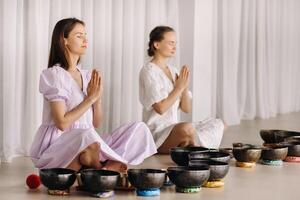 Two women are sitting with Tibetan bowls in the lotus position before a yoga class in the gym photo