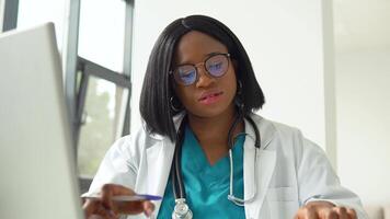 Young african american female student in white coat and stethoscope over her neck using laptop at desk while working at the university. Education concept video