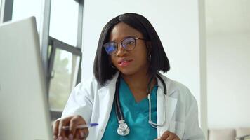 Attractive african american female doctor using laptop at desk while working in medical office video