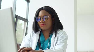 Young african american female student in white coat and stethoscope over her neck using laptop at desk while working at the university. Education concept video