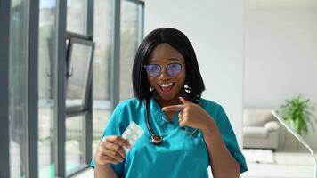 jovem africano americano mulher médico olhando às Câmera e mostra pílulas. sorridente fêmea médico posando dentro hospital escritório video