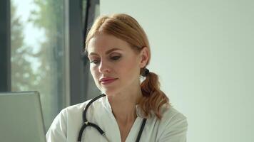 Attractive caucasian female doctor in white coat and stethoscope over her neck using laptop at desk, looking at camera while working in medical office video