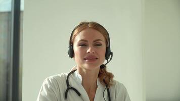 Professional female doctor in white medical coat and headset making conference call on laptop computer, consulting distance patient online in video chat