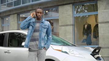 africano americano hombre hablando en el teléfono mientras cargando un eléctrico coche. ambientalmente simpático transporte video