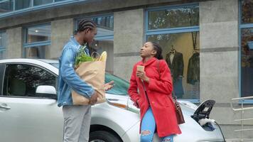 African american couple stands with groceries near electric car. Charging electric car at the electric gas station video