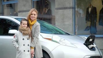 un mujer con su hija soportes cerca su eléctrico coche y mira a el cámara en el estacionamiento lote. cargando un eléctrico coche a un gas estación video