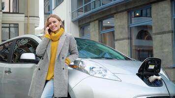 vrouw pratend Aan de telefoon terwijl opladen een elektrisch auto. milieuvriendelijk vriendelijk vervoer video