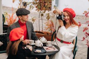 un elegante familia de Tres es sentado a un mesa fuera de en un café y Bebiendo café. papá, mamá y hija en el otoño ciudad foto