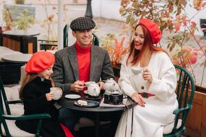 un elegante familia reunido juntos en un café en el calle. mamá, papá, pequeño hija bebida té, comer pasteles ellos son contento juntos. el concepto de un contento familia cena foto