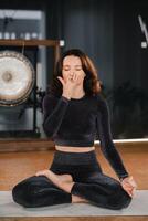 un mujer en un negro chandal se sienta en un yoga estera en un loto posición en el gimnasio foto