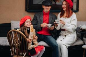 A stylish family of three is sitting at a table outside in a cafe and drinking coffee. Dad, mom and daughter in the autumn city photo