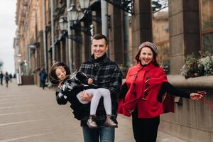 un elegante familia de Tres personas son caminando en el ciudad en el otoño y papá es participación su hija en su brazos foto