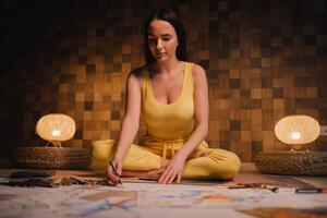 A girl in yellow clothes sitting in the Lotus position draws on paper with evening lighting at home photo