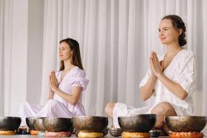 Two women are sitting with Tibetan bowls in the lotus position before a yoga class in the gym photo