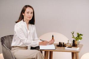 The aromatherapist girl is sitting in her office and holding a bottle of aromatic oil in her hands and writing something down. there are essential oils on the table photo