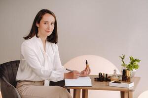 The aromatherapist girl is sitting in her office and holding a bottle of aromatic oil in her hands and writing something down. there are essential oils on the table photo