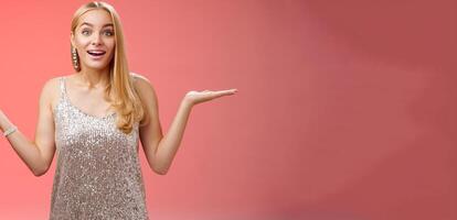 Carefree fascinated young blond girl 20s in silver glittering evening party dress raising hands sideways weighing shrugging smiling amused search right choice, deciding what do red background photo