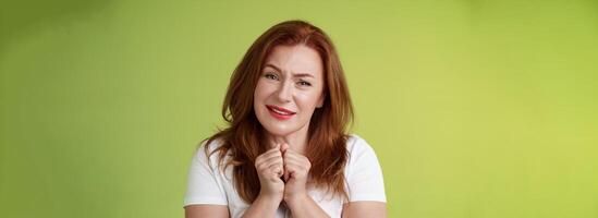 Silly touched tender redhead charmed middle-aged woman sighing gladly gaze admiration delighted press hands together heartwarmed fascinated look grateful lovely camera green background photo