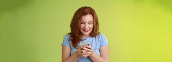 Intrigued excited tempting redhead middle-aged woman shopping online smartphone hold mobile phone look pleased entertained gadget screen smiling delighted playing game found perfect app photo