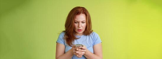 Confused unsure redhead middle-aged woman learn how use social media trying understand emoji look intense focused smartphone display reading important news message stand green background photo