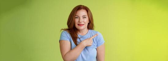 Surprised excited middle-aged wondered redhead woman pointing left amused standing thrilled joyful green background look camera curious interested cannot wait check-out great promo photo