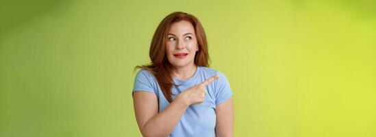 Excited intrigued middle-aged mature redhead woman pointing gazing left side copy space curiously smiling thrilled like interesting promo willing check-out great advertisement green background photo