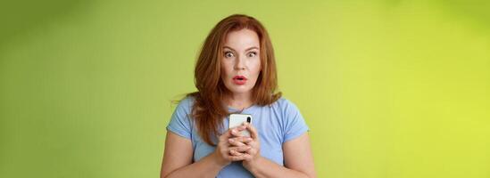 Shocked speechless impressed redhead european middle-aged woman fold lips astonished wow stare camera fascinated react amazed wondered received message hold smartphone intense green background photo