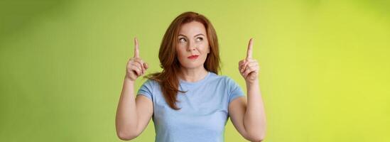 Curious thoughtful cute redhead female middle-aged ginger mother smirking thinking look aside pointing up index fingers deciding choice pondering choosing between variants green background photo