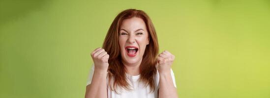 Yeah we did it. Joyful lucky redhead middle-aged female winner pump fists up celebration success gesture yelling triumph joy smiling broadly celebrate awesome news stand green background happy photo