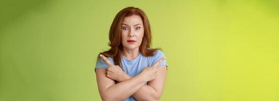 Help me choose. Worried unsure doubtful cute redhead middle-aged silly woman asking advice cross arms body pointing sideways left right look concerned camera make difficult decision green background photo