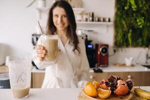 un contento sonriente sano mujer tiene preparado un Fruta cóctel y da eso a usted mientras en pie a hogar en el cocina.saludable comiendo foto