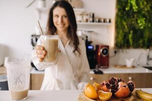 un contento sonriente sano mujer tiene preparado un Fruta cóctel y da eso a usted mientras en pie a hogar en el cocina.saludable comiendo foto