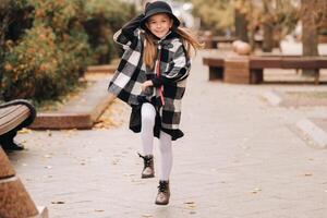 un elegante pequeño niña en un sombrero camina alrededor el otoño ciudad foto