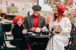 un elegante familia reunido juntos en un café en el calle. mamá, papá, pequeño hija bebida té, comer pasteles ellos son contento juntos. el concepto de un contento familia cena foto