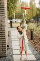 un pequeño niña con un cepillo limpia un camino en el calle en el patio foto