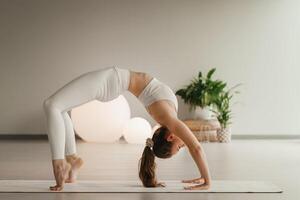 A girl in white clothes does yoga standing on the bridge on a mat indoors photo